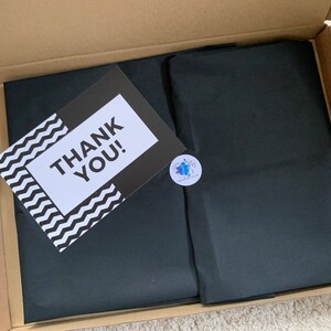 May include: A brown cardboard box with a black tissue paper liner. The tissue paper has a sticker with a blue and white design. A white card with black text that says "THANK YOU!" is on top of the tissue paper.