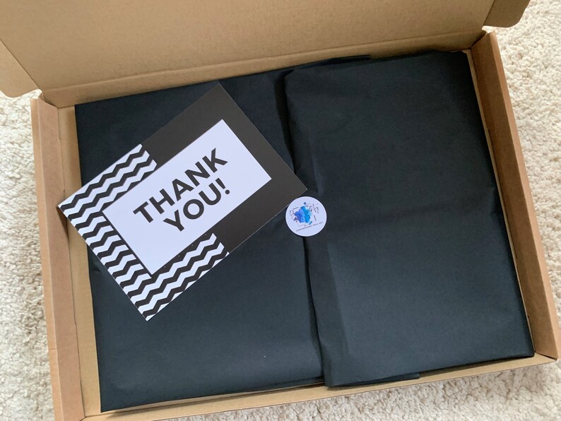 May include: A brown cardboard box with a black tissue paper liner. The tissue paper has a sticker with a blue and white design. A white card with black text that says "THANK YOU!" is on top of the tissue paper.