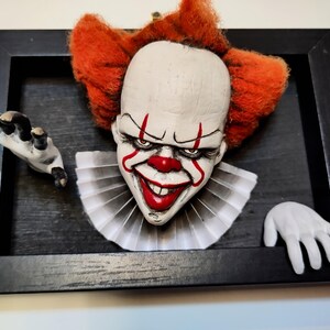 May include: A three-dimensional wall art piece featuring a clown head with bright orange hair, a white face with red makeup, and a pleated white collar. The clown is emerging from a black rectangular frame, with one hand reaching out.