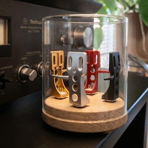 Könnte beinhalten: Eine transparente Glasglocke zeigt vier farbige Tonabnehmer: Gold, Silber, Rot und Schwarz. Die Tonabnehmer stehen auf einem Holzsockel. Im Hintergrund befindet sich ein Vintage-Stereoverstärker.