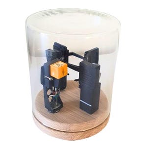 May include: A vintage turntable cartridge and stylus are displayed under a clear glass cloche. The cartridge has a bright orange component and is mounted on a wooden base. The cloche is cylindrical.