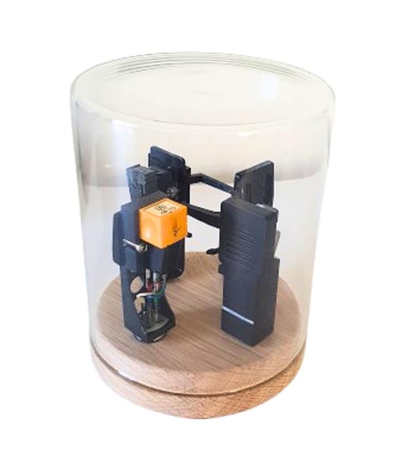 May include: A vintage turntable cartridge and stylus are displayed under a clear glass cloche. The cartridge has a bright orange component and is mounted on a wooden base. The cloche is cylindrical.