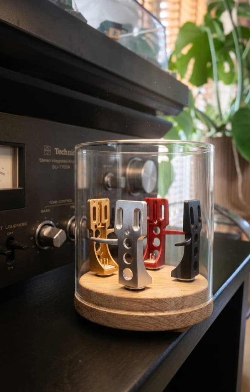 May include: A clear glass cloche displays four colored turntable headshells: gold, silver, red, and black. The headshells sit on a wooden base. A vintage stereo amplifier is in the background.