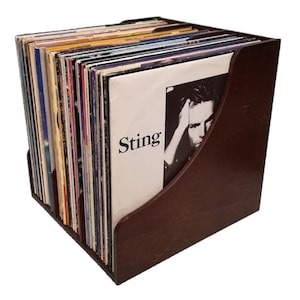 Op de afbeelding: Een donkerbruine houten platenhouder gevuld met vinylplaten. De albumhoes van Sting is zichtbaar. De houder heeft een vierkante vorm met gebogen zijkanten, ontworpen om vinylplaten op te bergen en weer te geven.