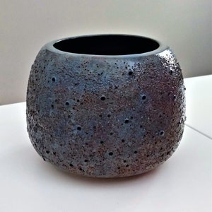 Può includere: Un vaso in ceramica blu scuro con una superficie testurizzata e piccoli fori. Il vaso ha una forma arrotondata e un'ampia apertura.