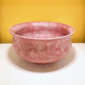 Pode incluir: Uma tigela grande de cerâmica rosa com uma textura marmorizada e com bolhas. A tigela é redonda e tem uma abertura larga e rasa.
