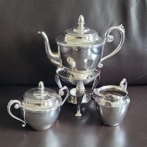 Op de afbeelding: Zilveren theeset met een theepot, roomkan en suikerpot. De theepot heeft een handvat en een tuit en staat op een standaard met een brander. De roomkan en de suikerpot hebben deksels.