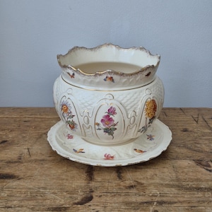 Op de afbeelding: Een crèmekleurige keramische plantenpot met een geschulpte rand en een bijpassende schotel. De buitenkant is voorzien van een gestructureerd vlechtpatroon, goudomrande boogpanelen en kleurrijke bloemenillustraties van rozen, madeliefjes en chrysanten.