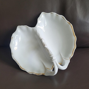 May include: White porcelain serving dish with a leaf-like design and a handle. The dish has a scalloped edge with gold trim. The handle is also white with gold accents. The dish is on a brown surface.