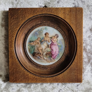 Op de afbeelding: Een ingelijst kunstwerk met een vintage porseleinen cameo met een geschilderd tafereel van drie vrouwen in een tuinsetting. De cameo is omgeven door een donkerhouten lijst.