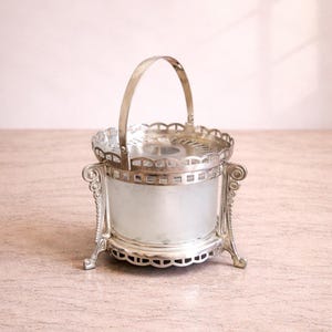 Könnte beinhalten: Antiker silberfarbener Eiskübel mit mattiertem Glas. Er hat einen dekorativen Griff, verzierte Beine und einen gewellten Rand. Das Design ist vintage und elegant, geeignet zum Servieren von kalten Getränken. Höhe ca. 15 cm.