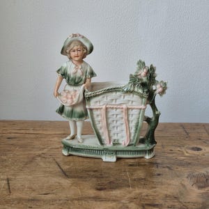 Op de afbeelding: Een porseleinen beeldje van een persoon in een groen-wit tenue met een mand fruit, naast een grote plantenbak in mandstijl versierd met roze linten en groen blad, geplaatst op een verhoogde ovale voet.
