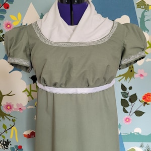 May include: A sage green empire line dress with short, puffed sleeves. The dress has white lace trim on the neckline, sleeves, and waist. A white shawl is draped over the shoulders. The dress is displayed on a dress form.