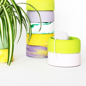 May include: A stack of six colorful ceramic planters in shades of green, yellow, and purple. The planters are stacked in a graduated size, with the largest planter at the bottom and the smallest planter at the top. A green plant is in the largest planter.