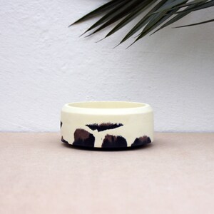May include: A white ceramic bowl with a black and brown abstract pattern. The bowl is round and has a smooth surface.
