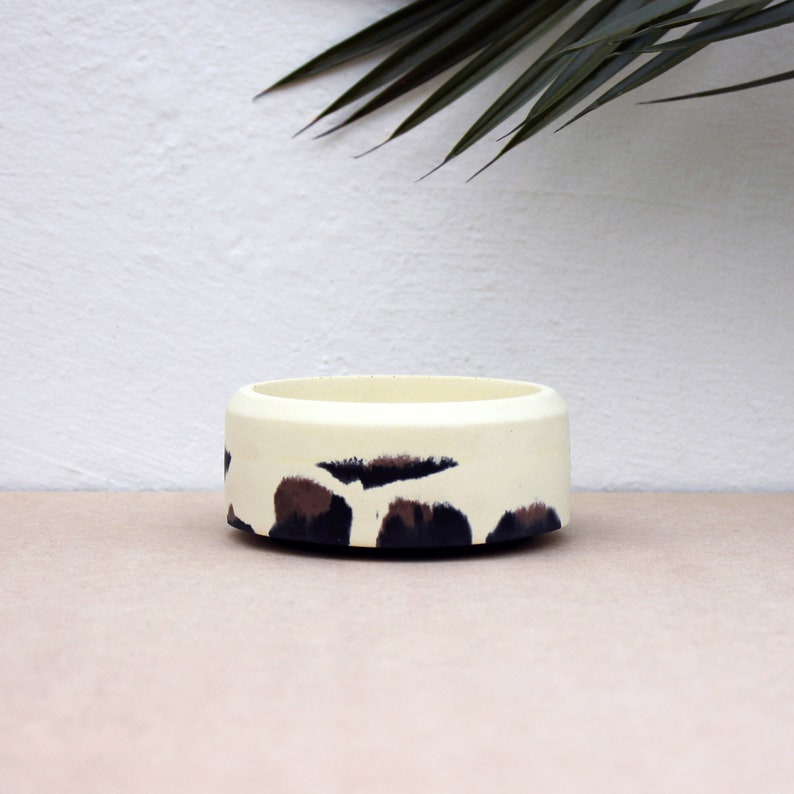 May include: A white ceramic bowl with a black and brown abstract pattern. The bowl is round and has a smooth surface.