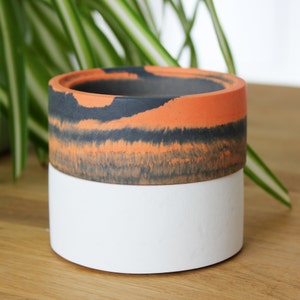 May include: A white and orange concrete planter with a black and orange marbled design. The planter is cylindrical and has a smooth finish.