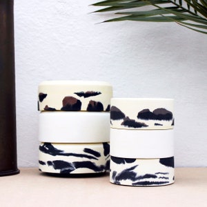 May include: Two sets of stacked white ceramic containers with black and brown abstract patterns. The containers are cylindrical and have a smooth, glossy finish.