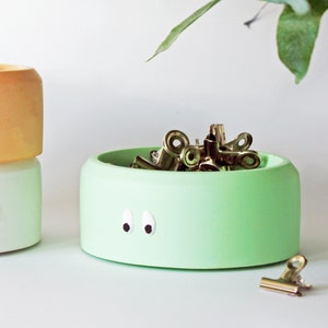 May include: A light green ceramic bowl with googly eyes, filled with gold binder clips. A single gold binder clip is on the surface next to the bowl.