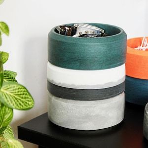 May include: A stacked set of four round concrete containers with a green, white, black, and gray finish. The top container has a watch and a leather band inside.