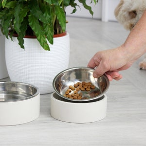 May include: Two white ceramic elevated dog bowls with stainless steel inserts. One bowl is filled with brown dog food.