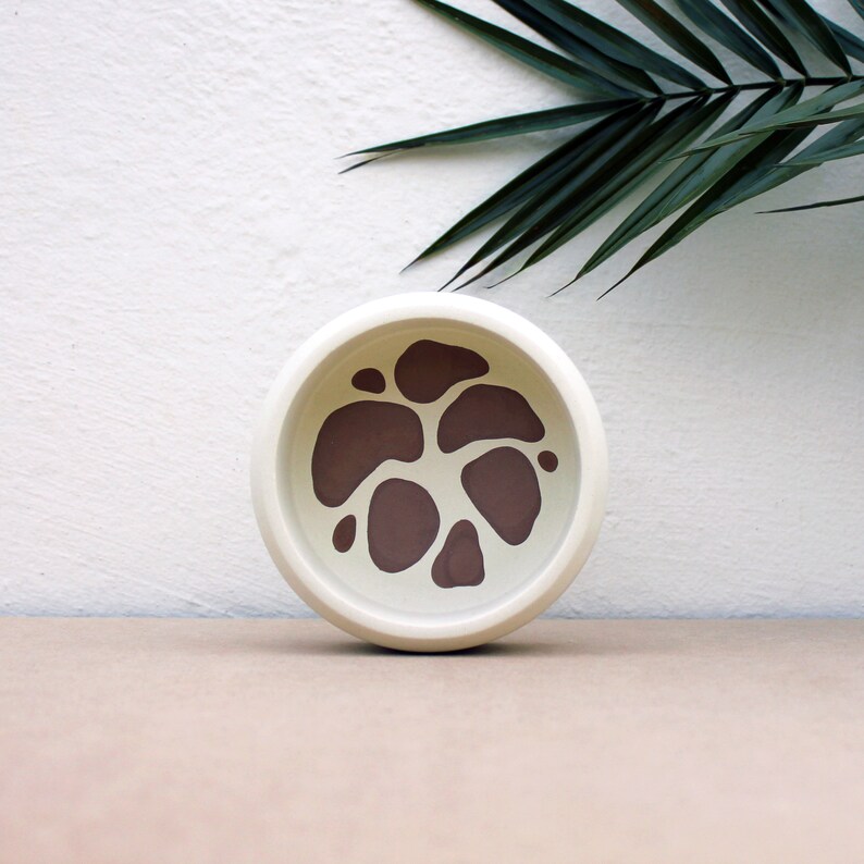 May include: A white ceramic pet bowl with a brown abstract design. The bowl is round and has a smooth, glossy finish.
