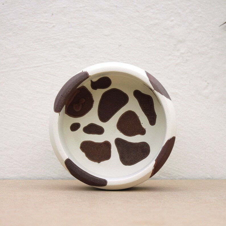 May include: A white ceramic bowl with a brown and white spotted pattern. The bowl is round and has a slightly raised rim.