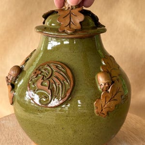 May include: A green ceramic jar with a lid decorated with oak leaves and acorns. The jar has a raised design of a dragon on the front.