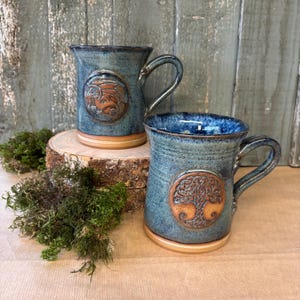 May include: Two blue ceramic mugs with handles, each with an embossed design. One features a dragon, the other a tree of life. Both have a light brown base and interior, displayed on a wooden slice with moss.