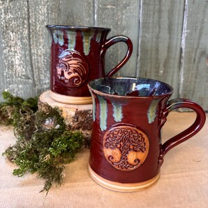 May include: Two burgundy ceramic mugs with a dragon and a tree of life design. The mugs have a dark green and light green glaze around the rim and down the sides. The mugs are on a wooden surface.