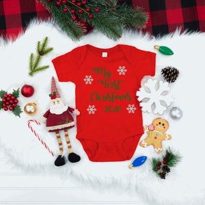 May include: Red baby onesie with the text "My First Christmas 2022" in green lettering and snowflakes. The onesie is surrounded by Christmas decorations, including a gingerbread man, a Santa figurine, and a candy cane.