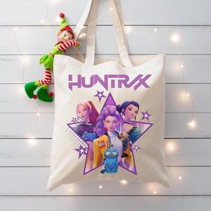 May include: A cream-colored tote bag featuring the word "HUNTRX" in purple, with a graphic of three women inside a purple star. A small blue cat is also featured. An elf doll is hanging on the side.
