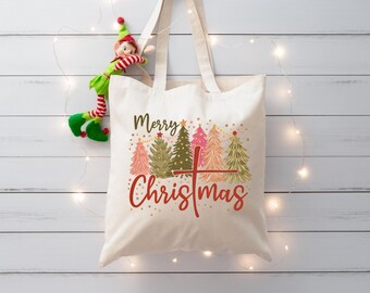 Puede incluir: Bolso tote color crema con las palabras "Merry Christmas" en escritura roja y marrón, con árboles de Navidad coloridos. Una muñeca elfo está sujeta a la bolsa. La bolsa tiene dos asas y cuelga sobre un fondo de madera blanca.