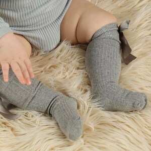 May include: A pair of gray ribbed knee-high socks with brown bows on the top. The socks are worn by a baby sitting on a fluffy white rug.