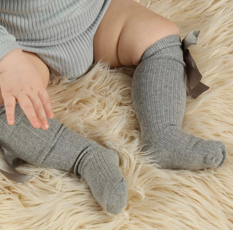 May include: A pair of gray ribbed knee-high socks with brown bows on the top. The socks are worn by a baby sitting on a fluffy white rug.