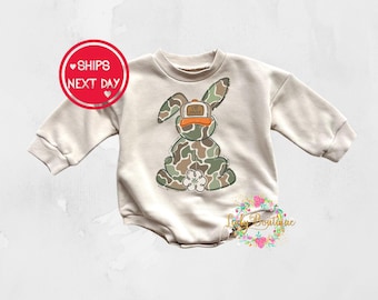 May include: A beige long-sleeved onesie with a camouflage bunny wearing a hat that says "Go Hunty".