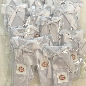 May include: A package of white knee-high socks with a white satin bow on each sock. The socks are packaged individually in clear plastic bags. The bags have a small white label with the text "Lilly Boutique" in black.