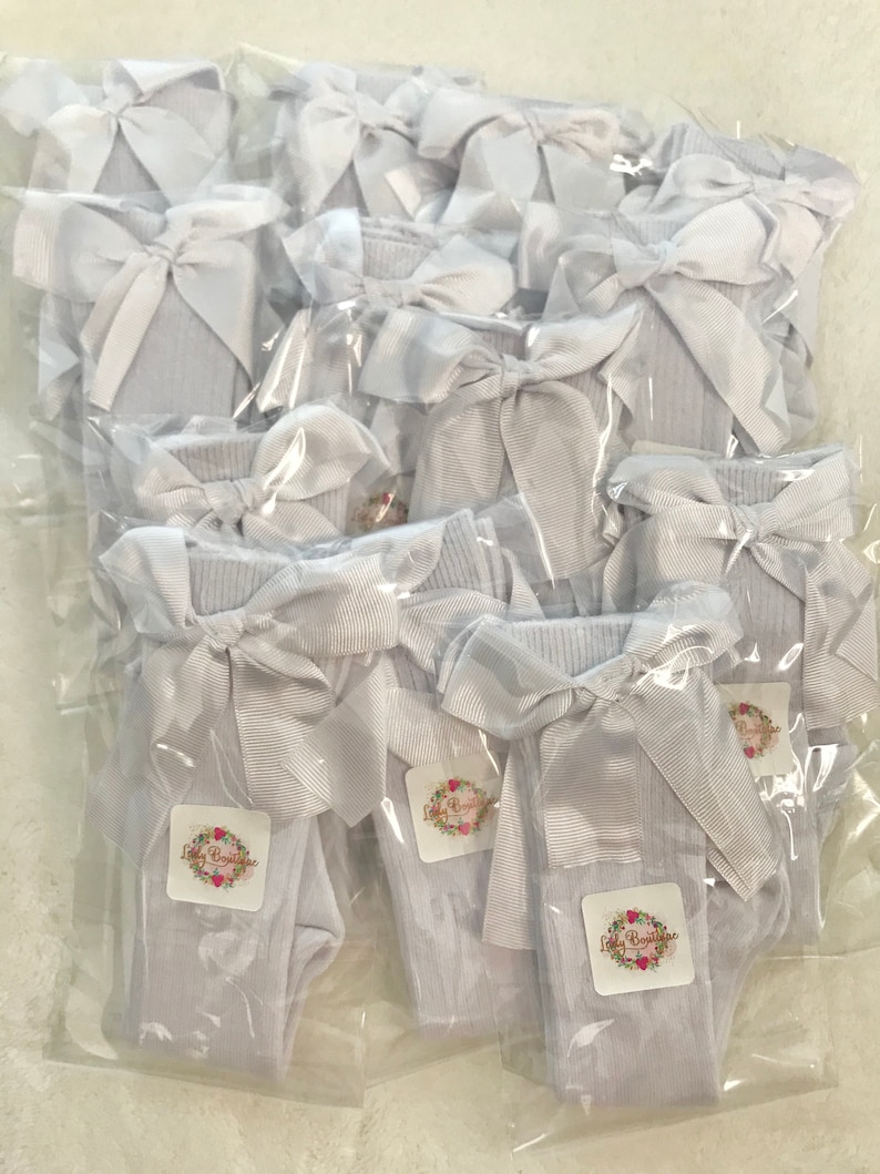 May include: A package of white knee-high socks with a white satin bow on each sock. The socks are packaged individually in clear plastic bags. The bags have a small white label with the text "Lilly Boutique" in black.
