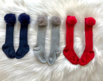 May include: Three pairs of knee-high socks with fuzzy pom-poms on top. The socks are in navy blue, gray, and red.