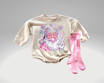 Puede incluir: Un mono beige de manga larga con un gráfico de Papá Noel rosa y blanco que lleva gafas de sol y sopla una burbuja. El mono se combina con calcetines rosas hasta la rodilla.