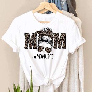 May include: White t-shirt with a leopard print design that spells out "MOM" with a woman's face in the middle. The woman has a messy bun and sunglasses. The text "#MOMLIFE" is below the design.