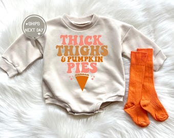 Puede incluir: Un body de bebé beige con el texto "Thick Thighs & Pumpkin Pies" en letras naranja, rosa y marrón. Un gráfico de pastel marrón y naranja se encuentra debajo del texto. Se incluyen dos pares de calcetines naranja hasta la rodilla.