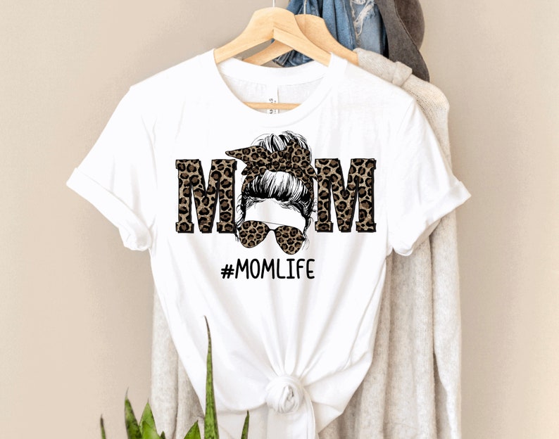 May include: White t-shirt with a leopard print design that spells out "MOM" with a woman's face in the middle. The woman has a messy bun and sunglasses. The text "#MOMLIFE" is below the design.