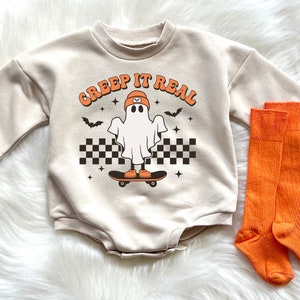 May include: A tan long-sleeve onesie with the text "Creep It Real" and a ghost wearing a hat skateboarding. The onesie is paired with a pair of orange knee-high socks.