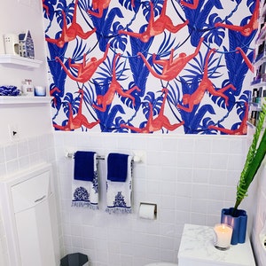 May include: A white bathroom with white tile walls and a white toilet. The bathroom has a white cabinet with a white door, a white sink with a chrome faucet, and a white countertop with a blue vase and a white candle. The walls are decorated with a wallpaper featuring red monkeys swinging on blue vines.