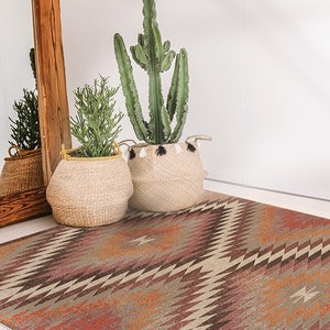 May include: A brown, beige and orange patterned rug with a geometric design. The rug is in front of a white wall and a wooden mirror. Two potted plants are in the background.