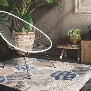 May include: A patterned rug with a geometric design in shades of blue, beige, and brown. The rug is made up of hexagonal tiles with different floral and geometric patterns. The rug is placed on a dark wood floor in a room with a white chair and a potted plant.