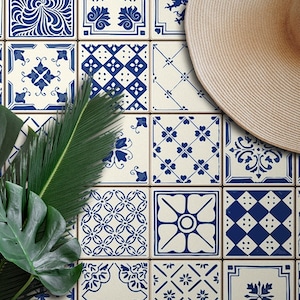 Op de afbeelding: Een verzameling witte keramische tegels met blauwe en witte patronen. De tegels zijn in een rasterpatroon gerangschikt en hebben verschillende geometrische en bloemmotieven. Een groene, bladerrijke plant en een strohoed liggen op de tegels.