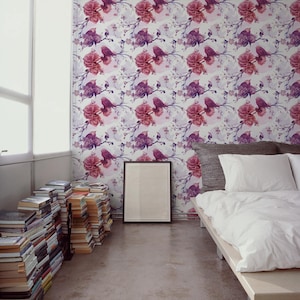 May include: A bedroom with floral wallpaper featuring pink and purple roses and birds. A bed with white linens and a wooden frame is in the foreground. Stacks of books line the wall, and a framed picture rests on the floor.