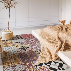 Puede incluir: Una alfombra de retazos con una variedad de patrones geométricos y florales en tonos de rojo, amarillo, azul y morado. La alfombra está colocada en un dormitorio, con una cama cubierta con una manta tejida de color beige. Una cesta tejida con un pequeño árbol está en el fondo.